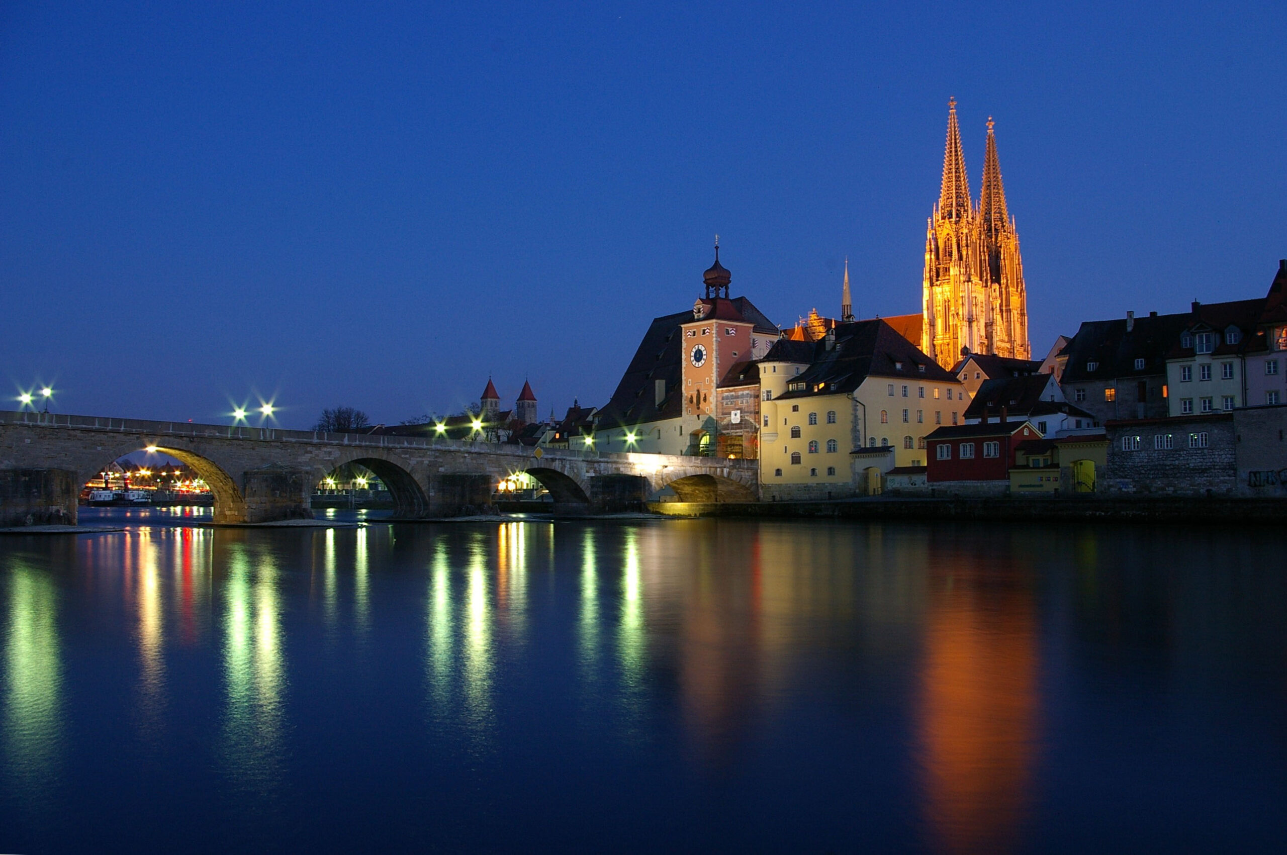 Eine Steinbrücke überspannt einen ruhigen Fluss in der Abenddämmerung, in dem sich die Gebäude der Stadt und eine hell erleuchtete Kathedrale unter einem tiefblauen Himmel im Wasser spiegeln.