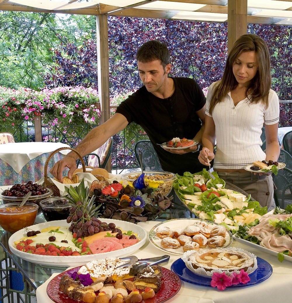 Ein Mann und eine Frau suchen sich an einem farbenfrohen Buffet mit Obst, Gebäck, Salaten und verschiedenen Gerichten, umgeben von Grünzeug und blühenden Blumen, etwas aus. Ein Mann und eine Frau suchen sich an einem farbenfrohen Buffet mit Obst, Gebäck, Salaten und verschiedenen Gerichten, umgeben von Grünzeug und blühenden Blumen, etwas aus.