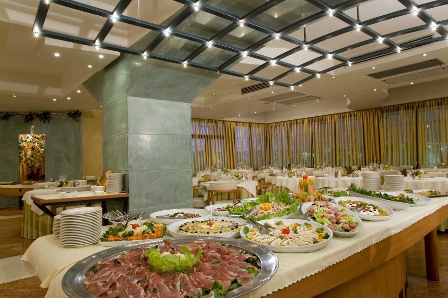 Ein Buffettisch in einem eleganten Restaurant ist mit verschiedenen Vorspeisen, Salaten und Aufschnittplatten gedeckt. Im Hintergrund stehen runde Tische mit weißen Tischtüchern und Stühlen, die durch große Fenster und drapierte Vorhänge geschützt sind. Ein Buffettisch in einem eleganten Restaurant ist mit verschiedenen Vorspeisen, Salaten und Aufschnittplatten gedeckt. Im Hintergrund stehen runde Tische mit weißen Tischtüchern und Stühlen, die durch große Fenster und drapierte Vorhänge geschützt sind.