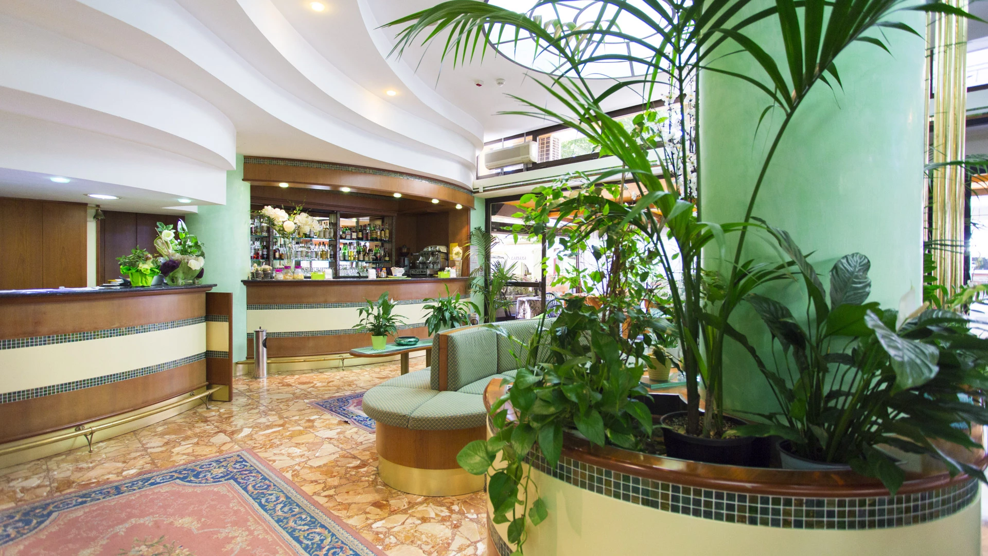 Eine Hotellobby mit grünen Säulen, großen Topfpflanzen, einem geschwungenen hölzernen Empfangstresen, einem Barbereich im Hintergrund, Marmorboden und bequemen Sitzgelegenheiten mit einem Teppich im Vordergrund. Eine Hotellobby mit grünen Säulen, großen Topfpflanzen, einem geschwungenen hölzernen Empfangstresen, einem Barbereich im Hintergrund, Marmorboden und bequemen Sitzgelegenheiten mit einem Teppich im Vordergrund.