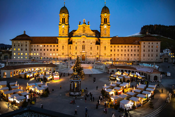 Ein großes, beleuchtetes Gebäude mit Zwillingstürmen überragt einen festlichen Weihnachtsmarkt in der Abenddämmerung. Lichterketten umgeben einen geschmückten Weihnachtsbaum in der Mitte des Platzes, um den herum die Menschen spazieren.