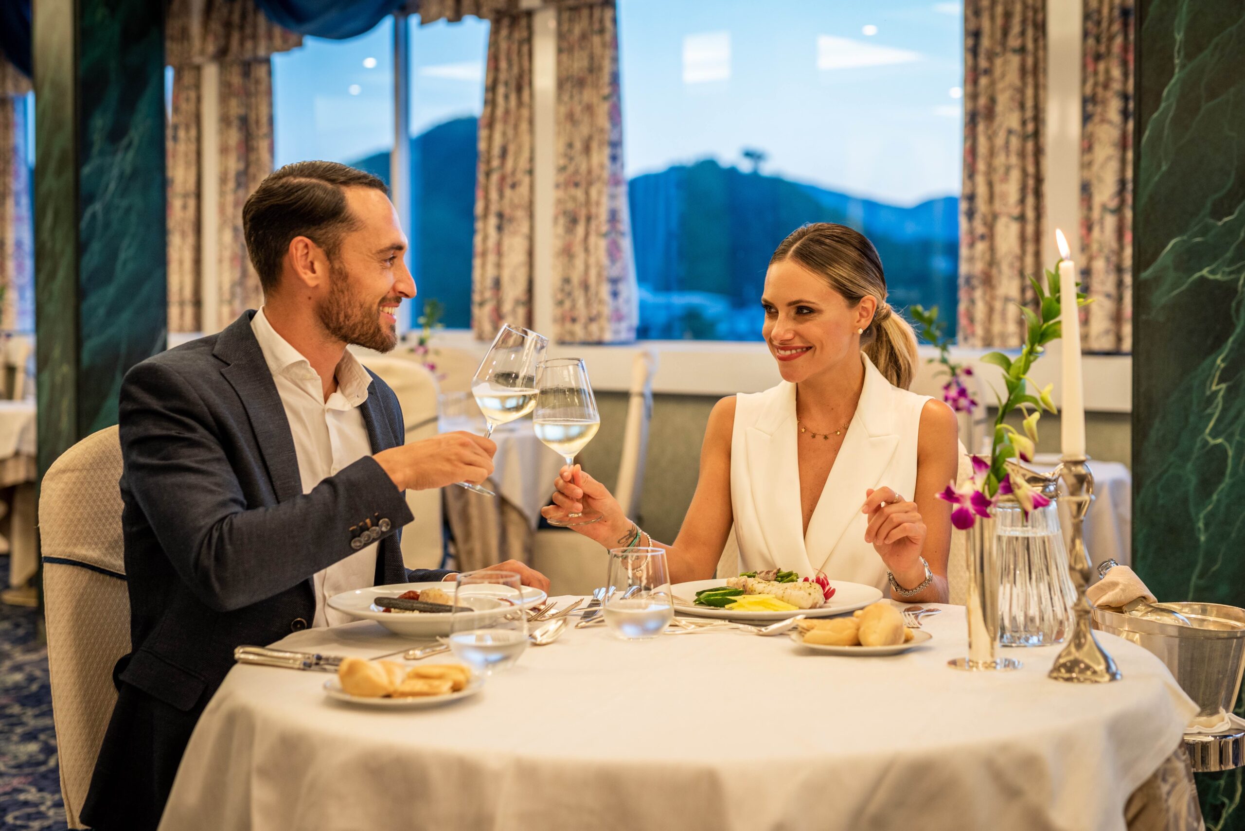 Ein Mann und eine elegant gekleidete Frau sitzen an einem Restauranttisch, lächeln und stoßen mit einem Glas Weißwein an. Ihr Tisch ist mit Speisen, Kerzen und Blumen gedeckt, was eine romantische und gehobene Atmosphäre schafft. Ein Mann und eine elegant gekleidete Frau sitzen an einem Restauranttisch, lächeln und stoßen mit einem Glas Weißwein an. Ihr Tisch ist mit Speisen, Kerzen und Blumen gedeckt, was eine romantische und gehobene Atmosphäre schafft.