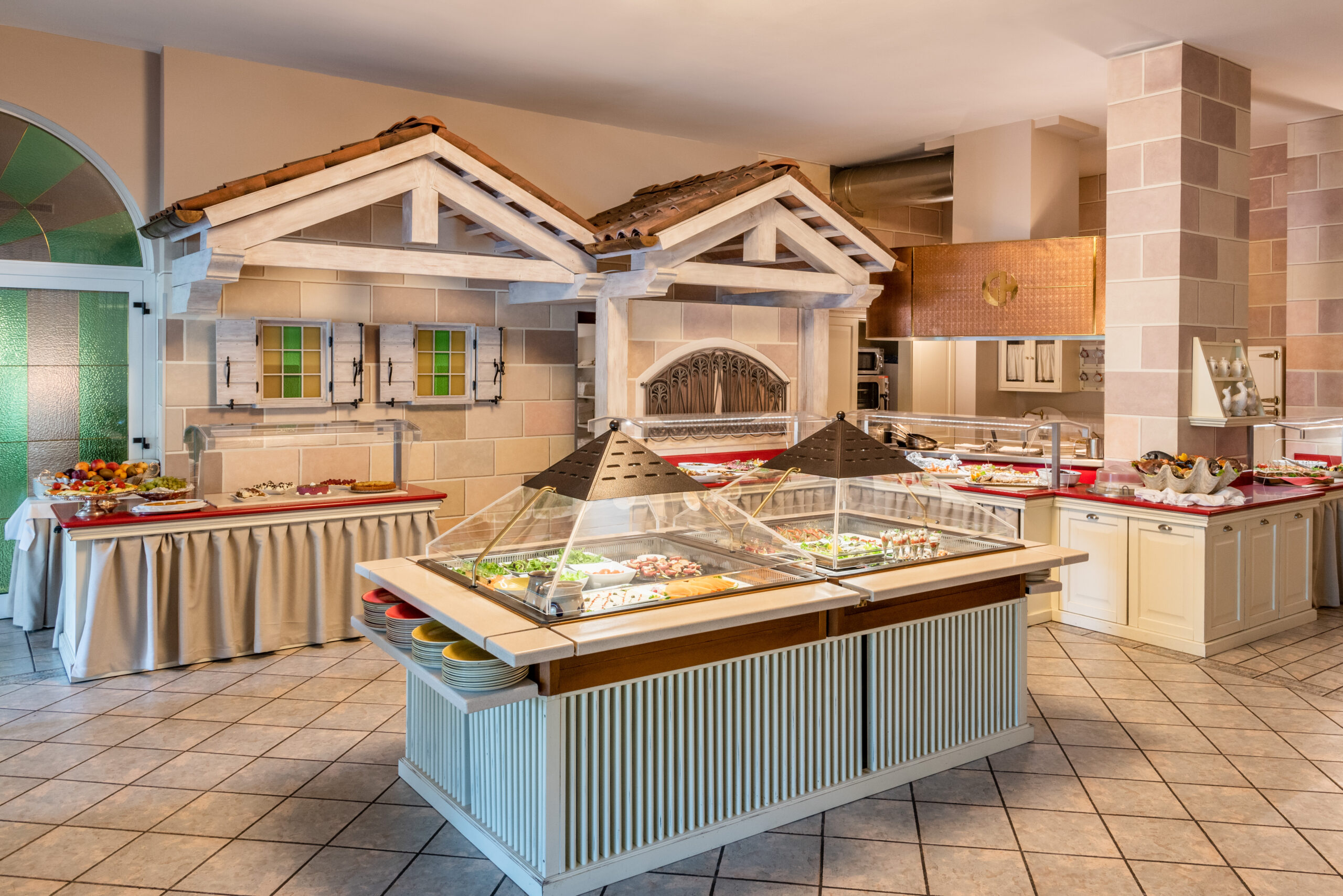 Ein Buffetrestaurant mit gefliestem Boden und dekorativen Strukturen, die an kleine Häuser erinnern, an der Rückwand. Salatbars und Tabletts sind ordentlich auf den Theken angeordnet, so dass eine Vielzahl von Gerichten sichtbar ist. Ein Buffetrestaurant mit gefliestem Boden und dekorativen Strukturen, die an kleine Häuser erinnern, an der Rückwand. Salatbars und Tabletts sind ordentlich auf den Theken angeordnet, so dass eine Vielzahl von Gerichten sichtbar ist.