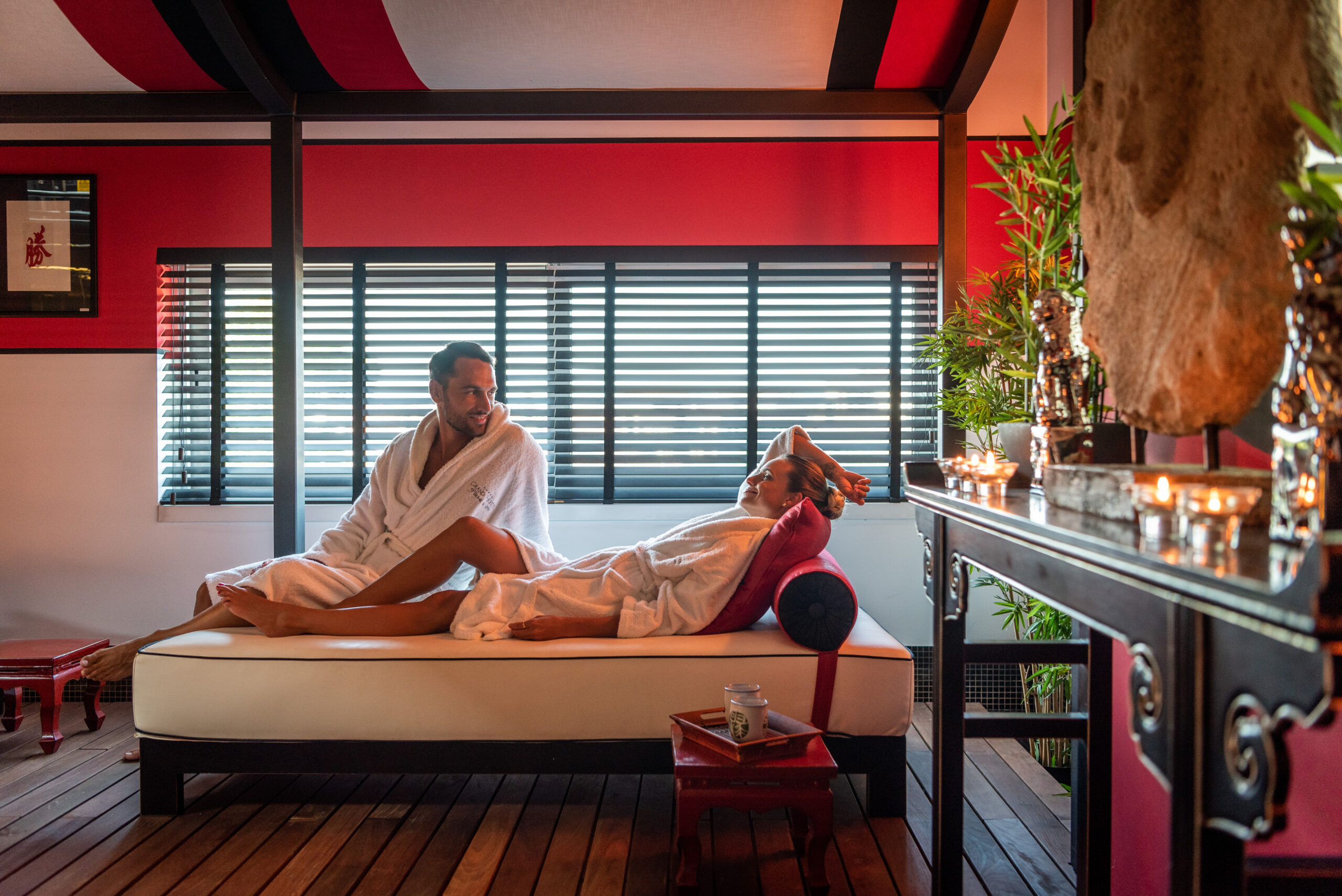 Ein Mann und eine Frau in weißen Gewändern entspannen sich auf einem Liegestuhl in einem mit Kerzen, Pflanzen und roten Akzenten dekorierten Wellnessbereich. Das Paar scheint zu lächeln und einen friedlichen Moment miteinander zu genießen. Ein Mann und eine Frau in weißen Gewändern entspannen sich auf einem Liegestuhl in einem mit Kerzen, Pflanzen und roten Akzenten dekorierten Wellnessbereich. Das Paar scheint zu lächeln und einen friedlichen Moment miteinander zu genießen.