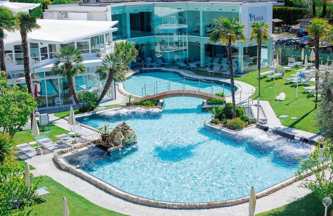 Ein luxuriöser Swimmingpool im Freien mit einer kleinen Holzbrücke, umgeben von Palmen, Liegestühlen, Sonnenschirmen und modernen Glasgebäuden mit der Aufschrift "Plaza" im Hintergrund. Üppige grüne Rasenflächen umrahmen den Poolbereich.