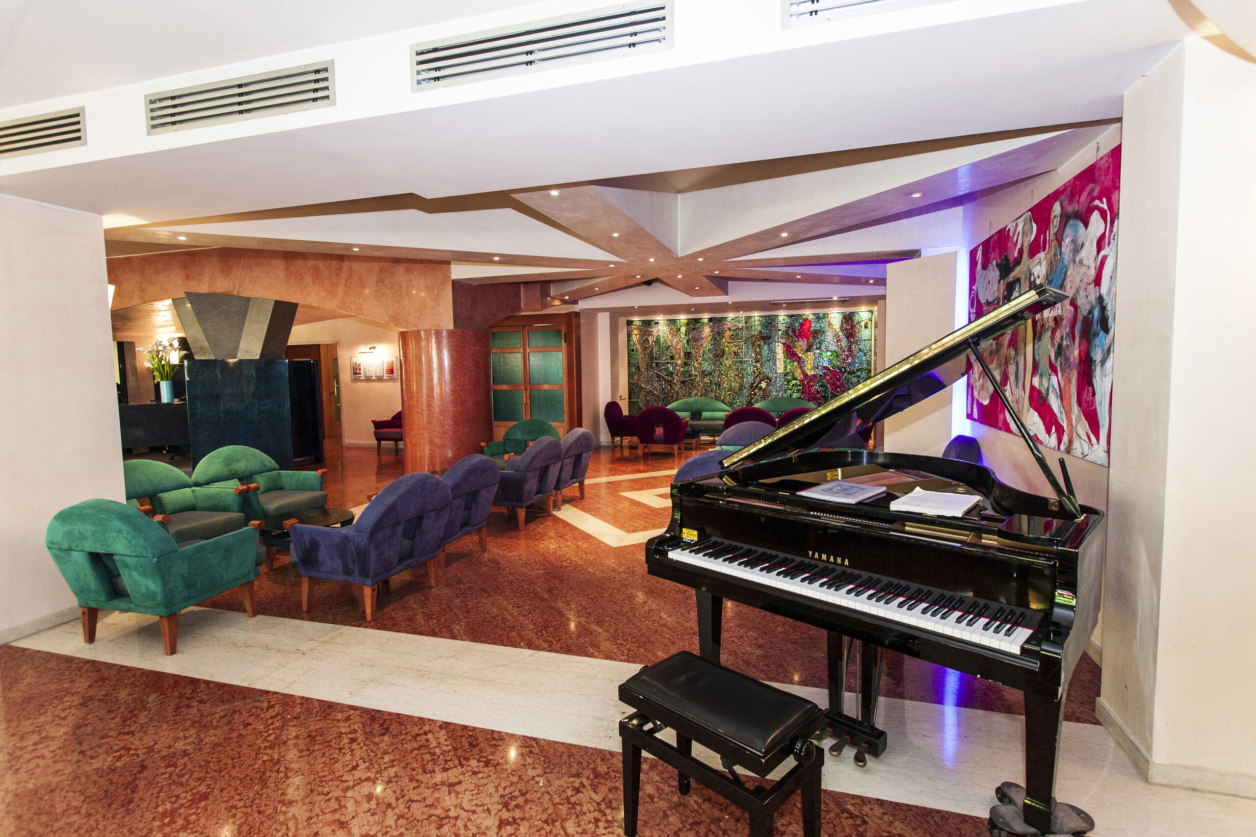 Eine Hotellobby mit grünen Sesseln, einem Flügel mit Bank, lebhaften abstrakten Kunstwerken an den Wänden und warmer Beleuchtung, die eine einladende Atmosphäre schafft. Eine Hotellobby mit grünen Sesseln, einem Flügel mit Bank, lebhaften abstrakten Kunstwerken an den Wänden und warmer Beleuchtung, die eine einladende Atmosphäre schafft.